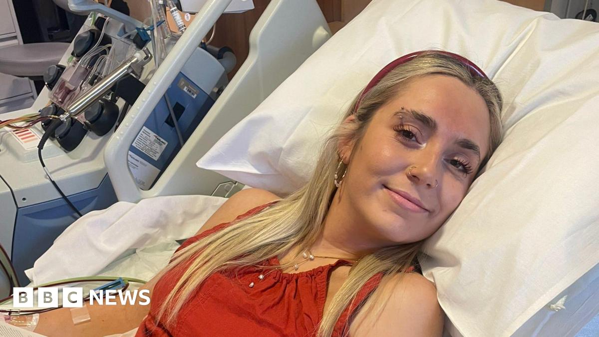 Beth Bowyer is laying down in a hospital bed and is wearing a red top and headband. She's smiling at the camera.