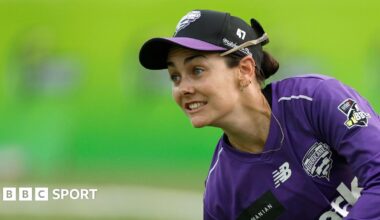 Heather Graham in action wearing a purple long sleeve kit and a black cap with a purple peak. Her sunglasses are on the back of her cap and she has a long gold chain around her neck.