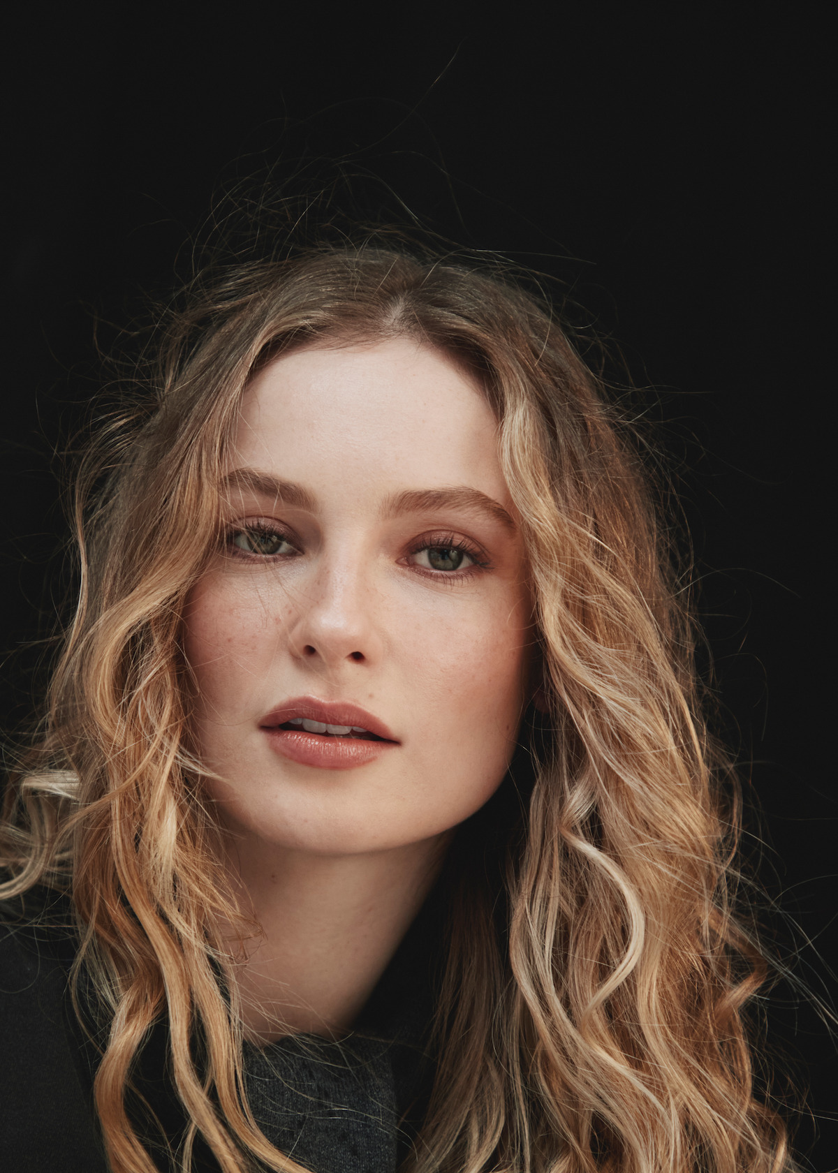 Portrait of a woman with long, wavy blonde hair and light makeup, looking directly at the camera against a plain dark background in a studio setting.