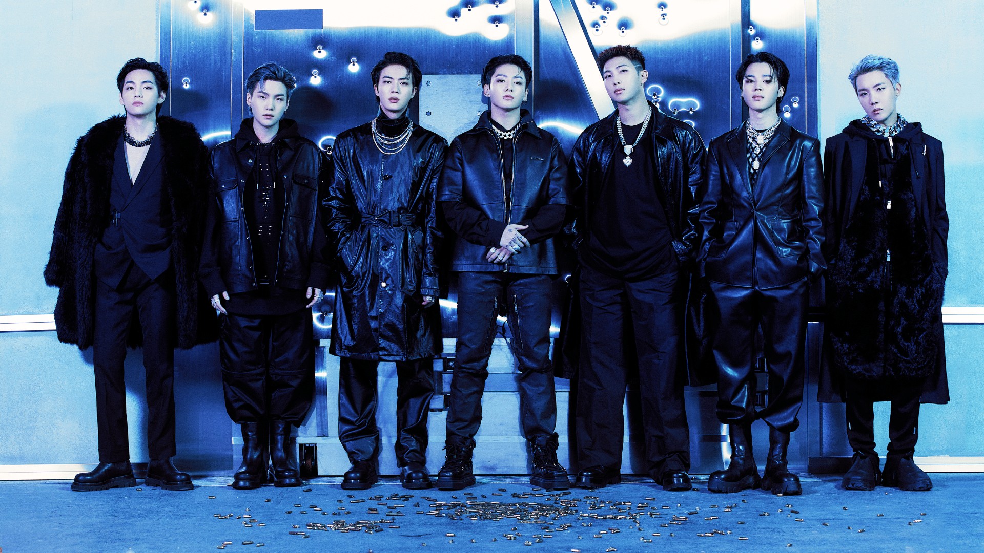 Seven members of BTS dressed in black leather and fur outfits stand in a line against a blue-lit, metallic vault background marked with bullet holes.