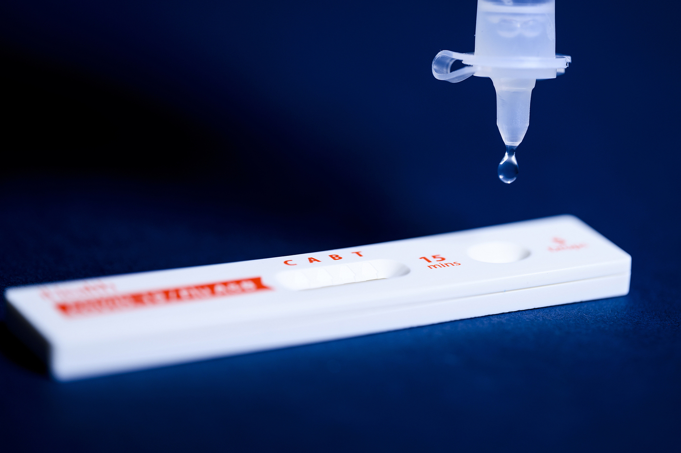 A COVID-19 test lying flat with a droplet falling from a syringe.