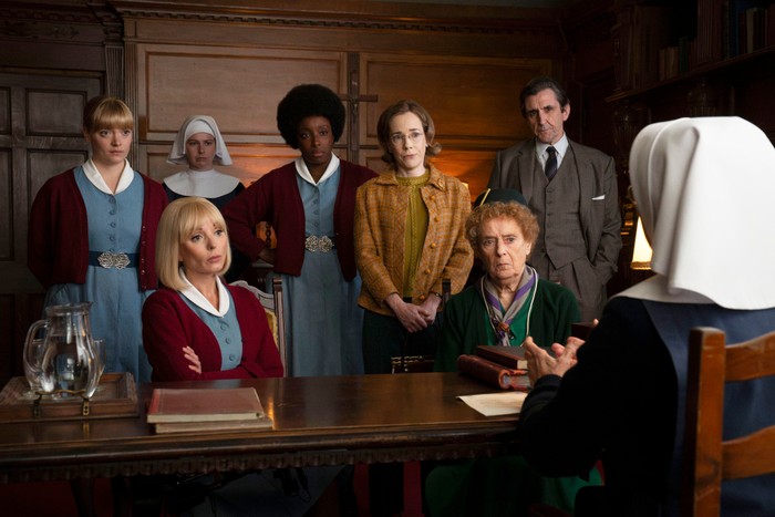 A group of midwives, nuns and colleagues stand and sit around a wooden table in a wood-panelled room, listening intently as a nun speaks to them.