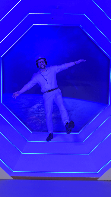 Man posing playfully with arms outstretched inside a blue neon-lit hexagonal tunnel installation, wearing a helmet and standing against a space-themed backdrop.