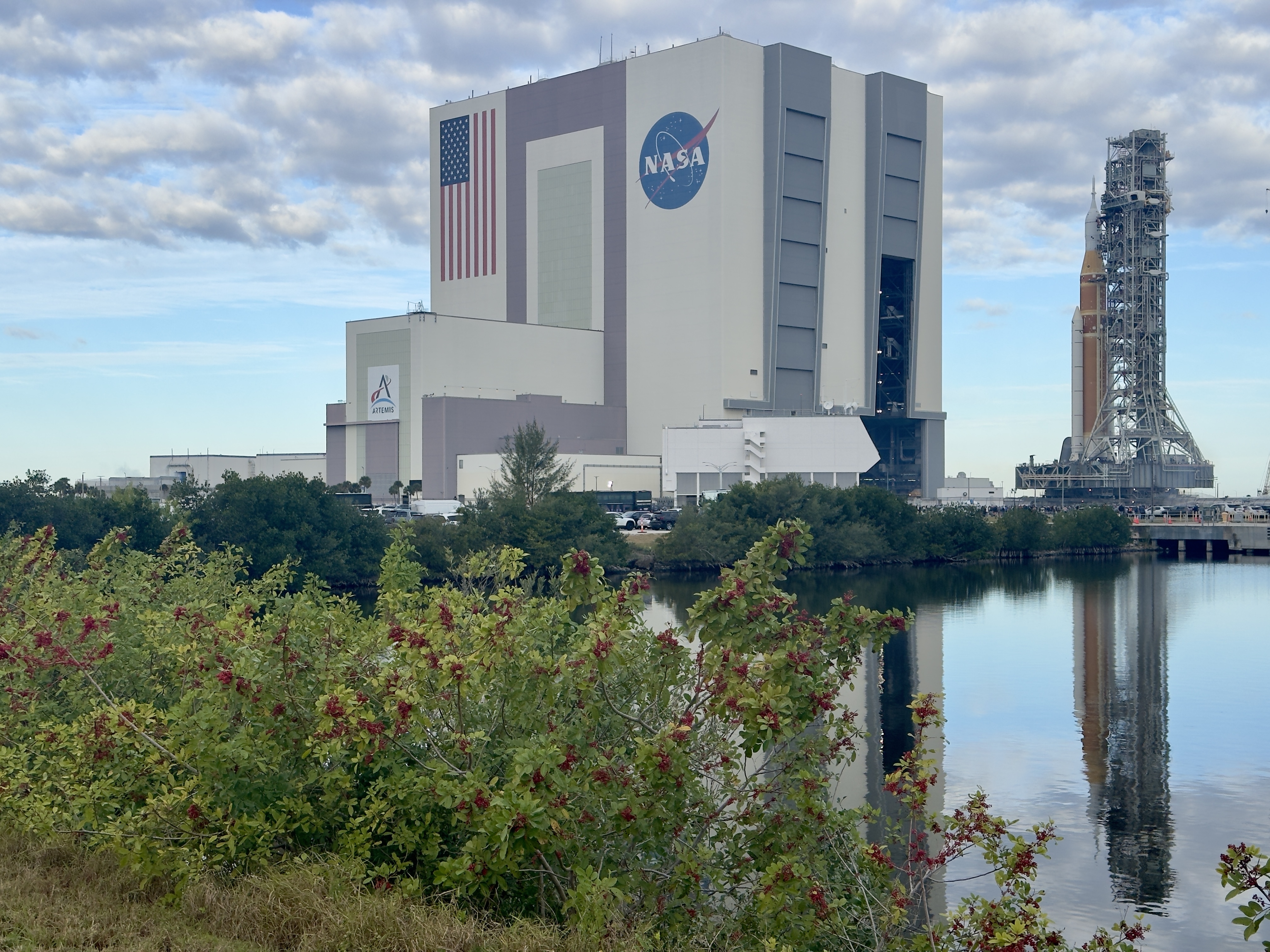 A giant rocket rolls out to toward the launch pad and is reflected in a lagoon for artemis 2