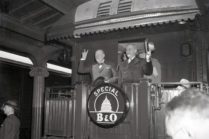 Winston Churchill and US president Harry S Truman stand on the rear platform of a train, both raising “V for Victory” signs to a crowd in 1946.
