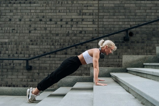 Beautiful athletic blonde woman in sportswear doing push-ups on the stairs from a step on the city street, warming up before jogging. Sport concept.