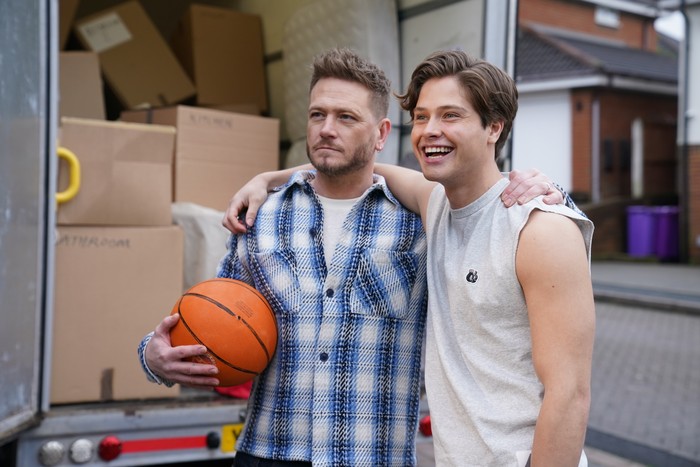 Matthew Wolfenden, holding a basketball, with his arm around Lewis Walton filming a scene for Hollyoaks beside a removals van