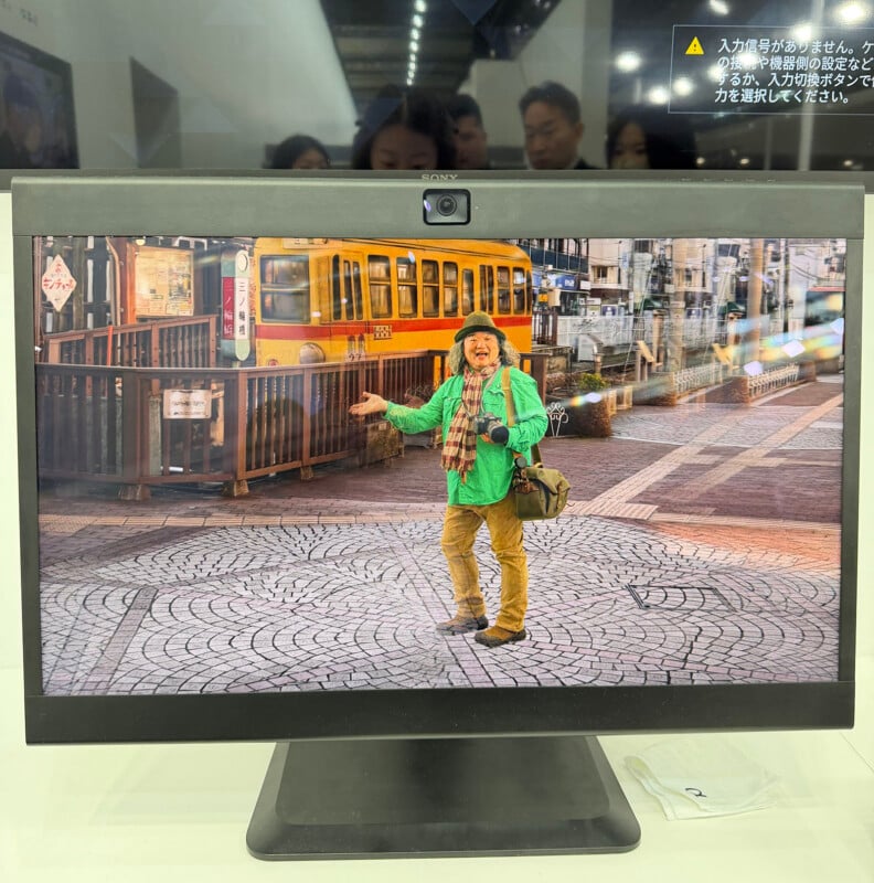 A computer monitor displays an image of a cheerful man in colorful clothing and a hat, standing on a cobblestone street with a yellow tram and buildings in the background. Reflections of people can be seen above the monitor.