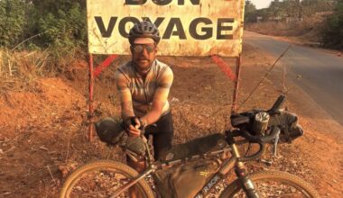 Swiss cyclist caked in dust standing in front of a sign in the Republic of Guinea