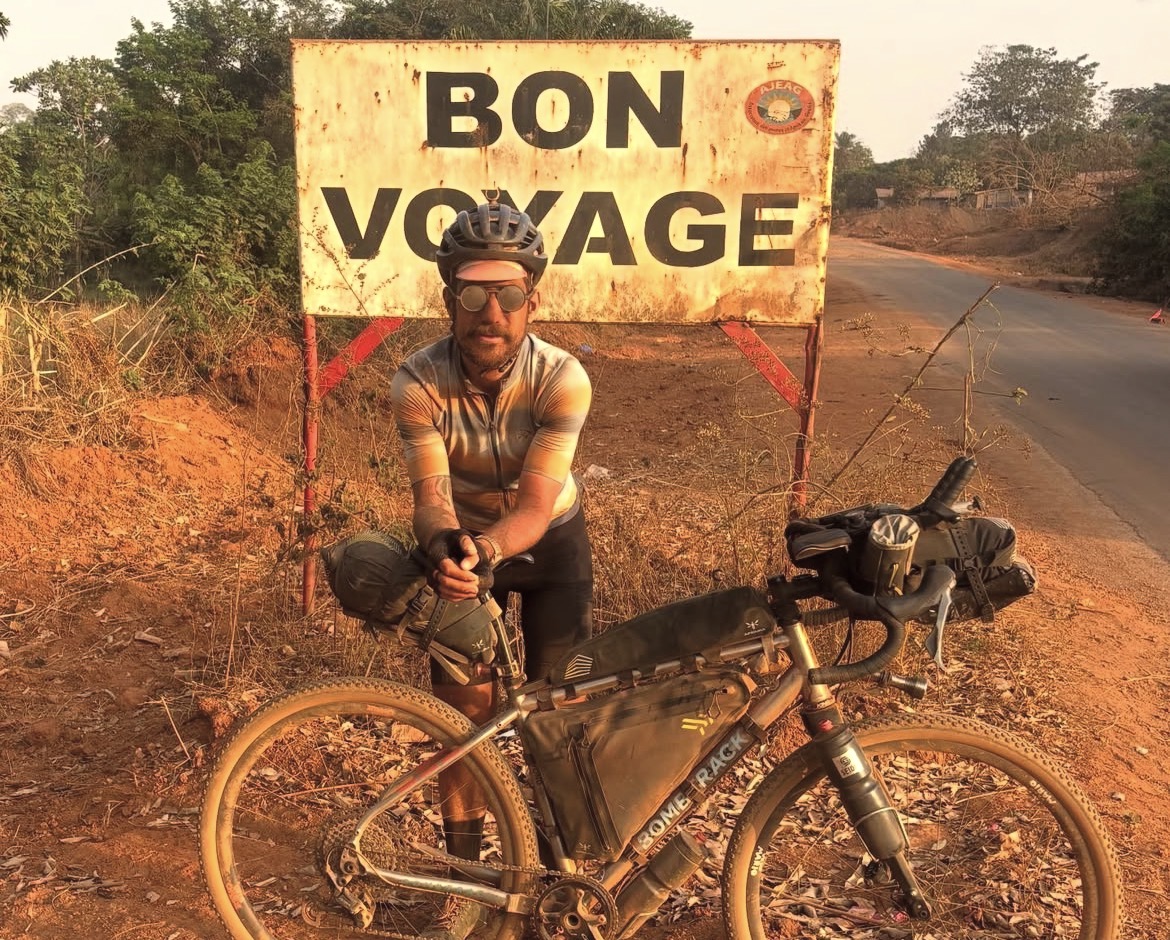 Swiss cyclist caked in dust standing in front of a sign in the Republic of Guinea