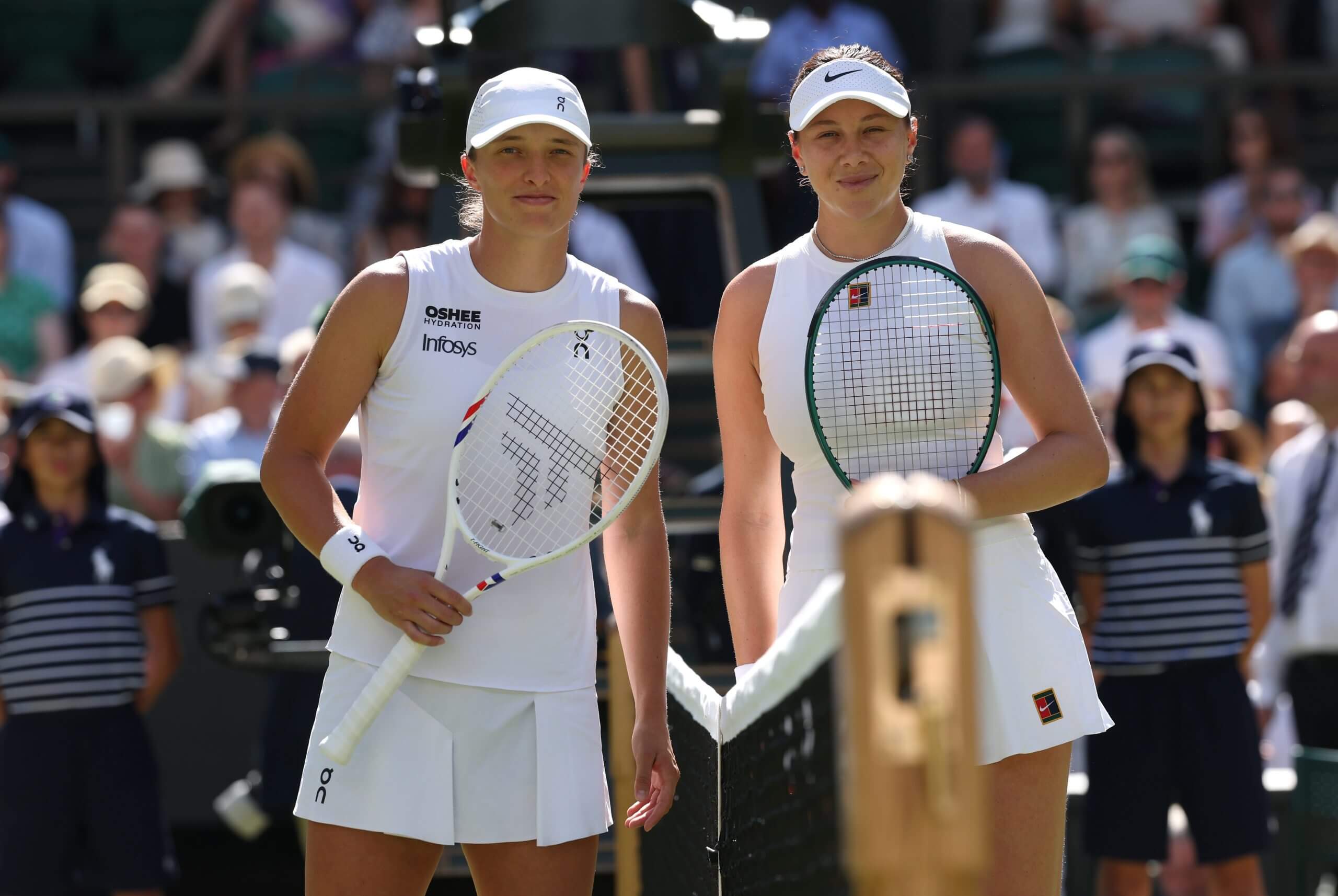 Iga Świątek and Amanda Anisimova stand together on either side of a tennis net with a crowd behind them.