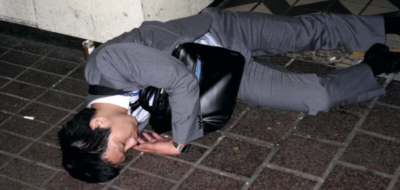 A man in a gray suit is lying on his side on a tiled sidewalk, appearing to be asleep. He has a black bag across his chest, and a can is visible near his head.
