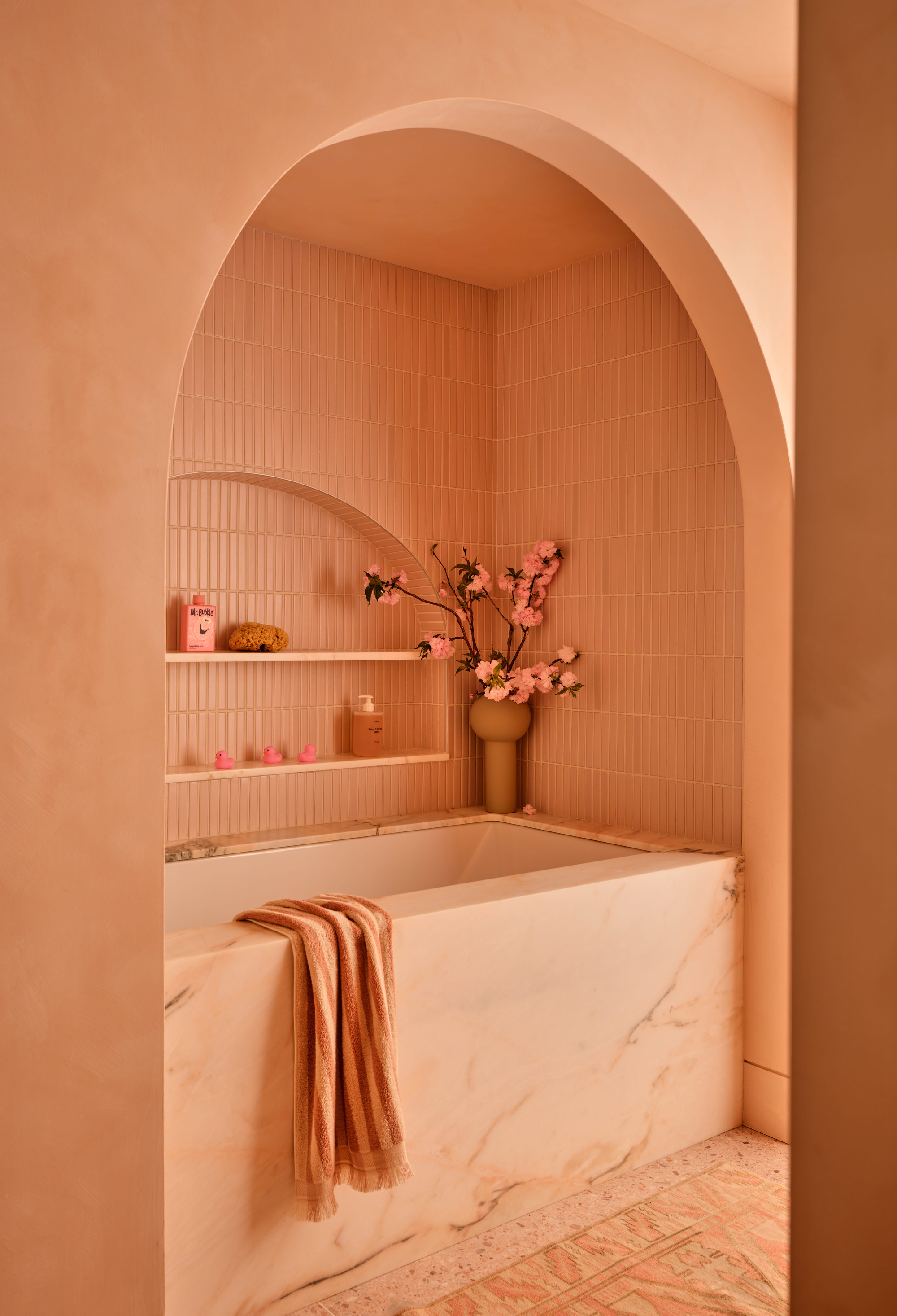 Image of a marble bathtub in an alcove with an arched doorway. The alcove is covered in subway tiles, and there is a towel draped over the tub. Everything is pink.