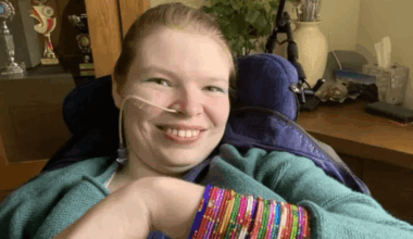 Lucinda Ritchie, smiling, in her wheelchair in a domestic setting, with a small breathing tube coming from her nose