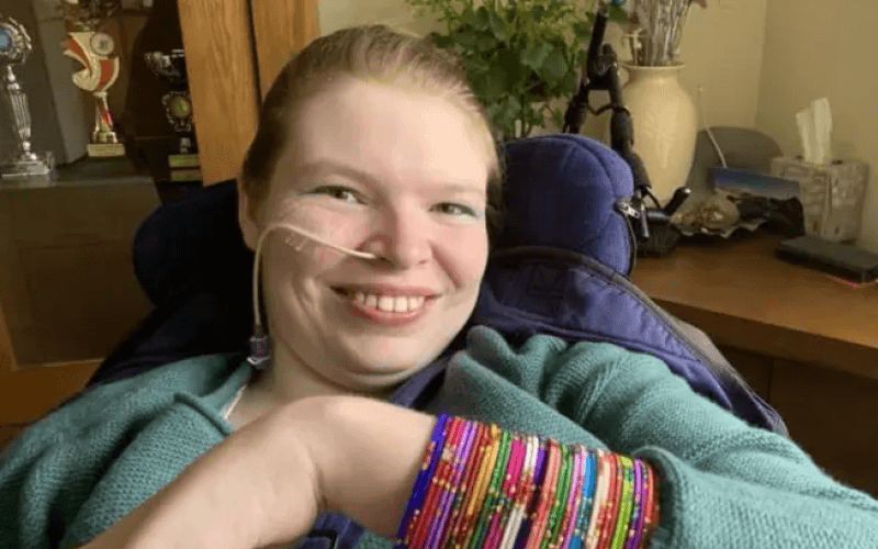 Lucinda Ritchie, smiling, in her wheelchair in a domestic setting, with a small breathing tube coming from her nose