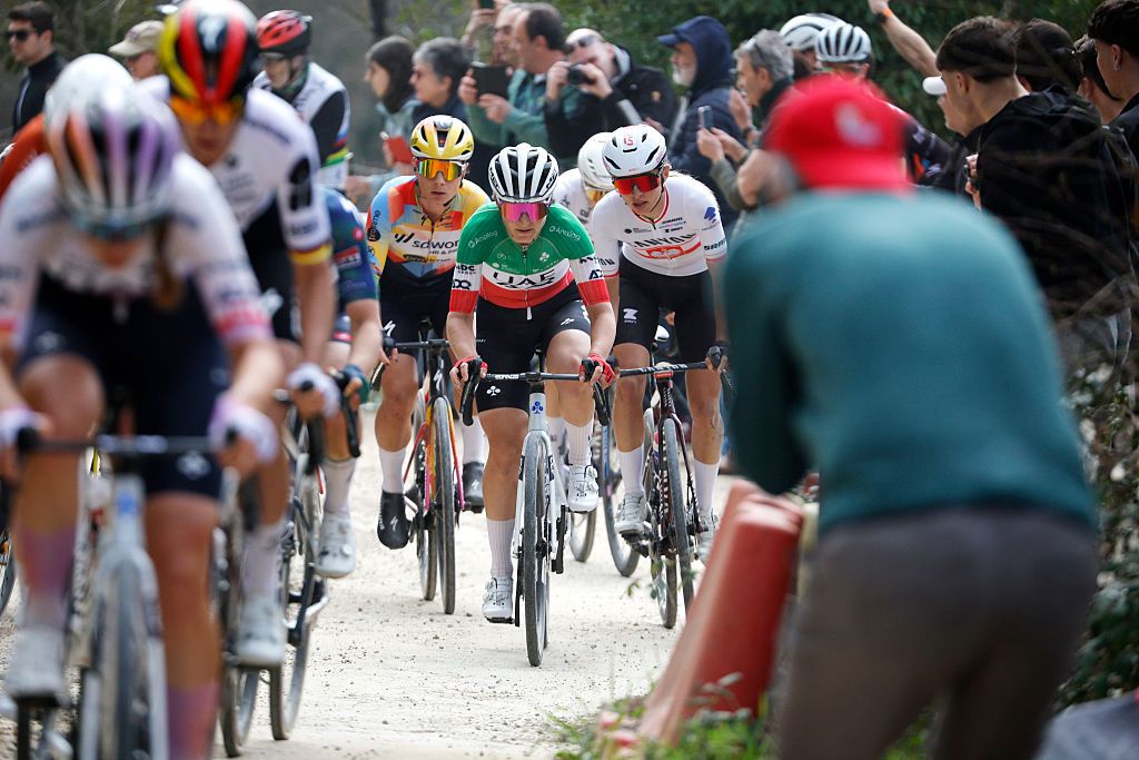 A long shot, taken at Strade Bianche Women 2026, showing blurred riders in the foreground and then focusing in on Elisa Longo Borghini, Lotte Kopecky and Kasia Niewiadoma-Phinney behind