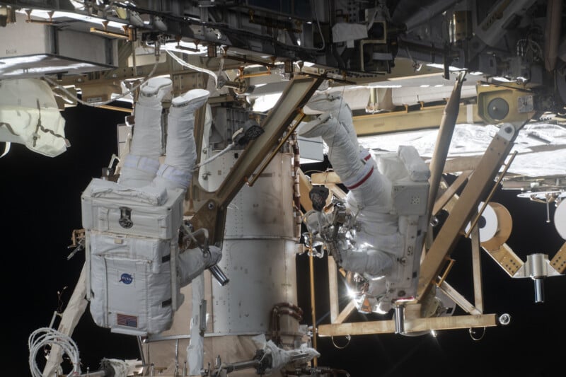 Two astronauts in white spacesuits work outside the International Space Station, attached to metal framework and equipment, with black space in the background.