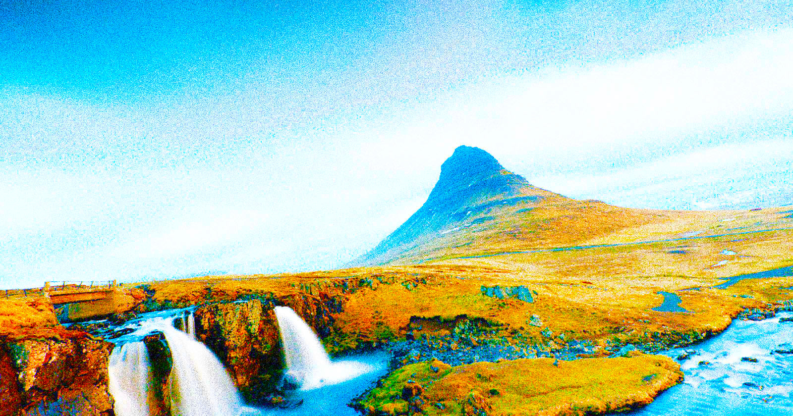 A grassy landscape with a small waterfall in the foreground and a distinctively shaped mountain under a bright, mostly clear sky.