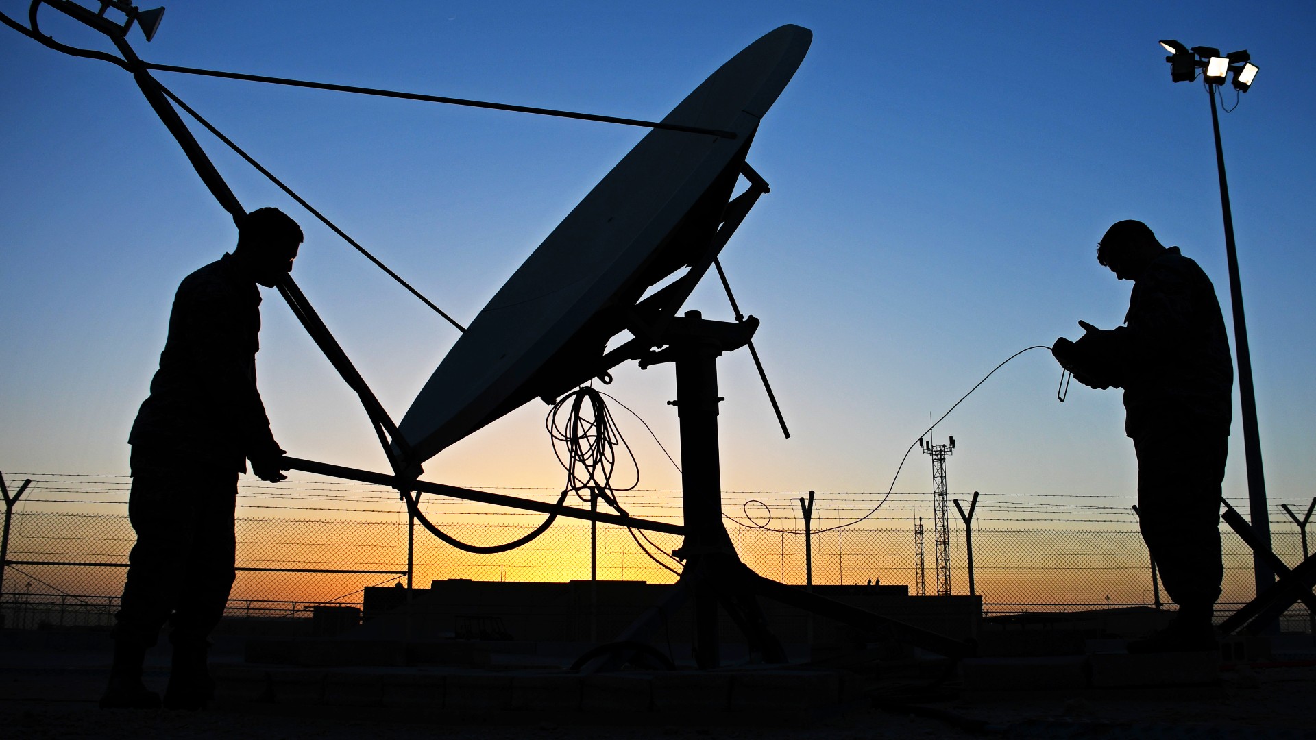 silhouettes of two people beside a large antenna dish at sunset, next to chain link fences topped with barbed wire