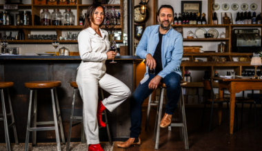 A woman in a white jumpsuit and red boots stands with a wine glass, while a man in a blue shirt and jeans sits on a bar stool holding a glass.