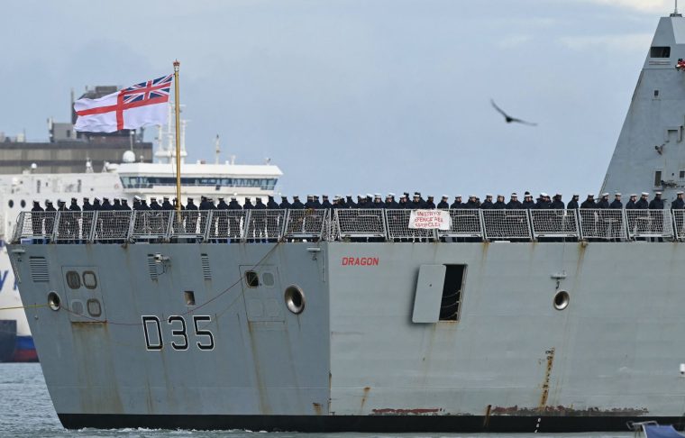 (FILES) Crew members stand beneath an Ensign on the aft deck of HMS Dragon, a Royal Navy Type 45 Daring-class air-defence destroyer warship, as it is guided by tug boats from HM Naval Base Portsmouth, on the south coast of England, on March 10, 2026. The Middle East war, launched on 28 February, is exposing the limits of Britain's navy, seen as humiliating for a country long considered a historic maritime power. Controversy about Britain's naval capabilities first erupted over the late deployment of HMS Dragon to protect British bases in Cyprus, one of which was hit by an Iranian drone. The destroyer has only just arrived. In the meantime, France deployed its Charles de Gaulle aircraft carrier, while Italy and Greece sent ships to reassure Cyprus. Right-wing newspaper the Daily Telegraph called the comparisons "embarrassing". (Photo by JUSTIN TALLIS / AFP via Getty Images)