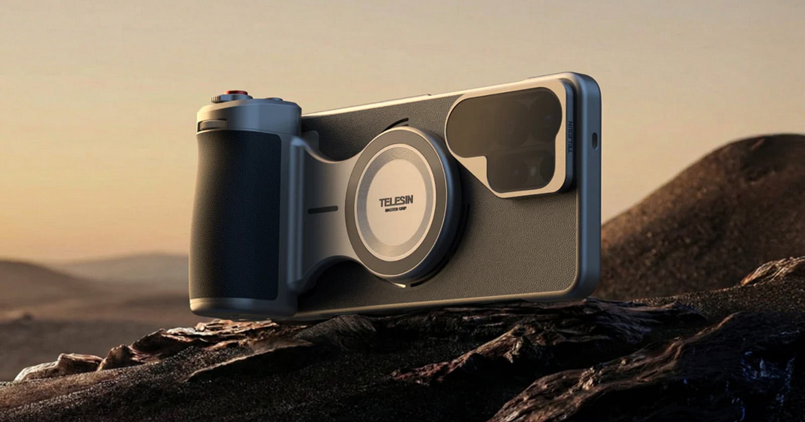 A smartphone with a large camera and ergonomic grip attachment rests on a rocky surface with a desert landscape in the background during sunset.