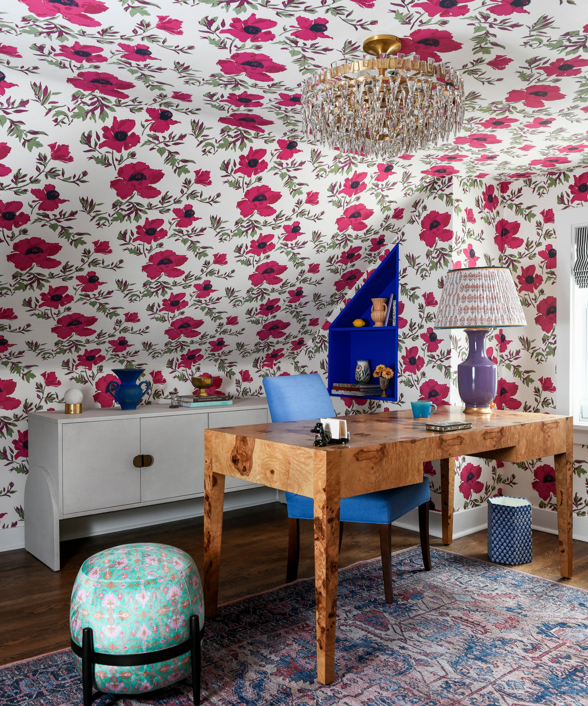 A home office with hot pink floral wallpaper on the walls and a sloped ceiling, a burl wood desk, a purple and blue rug, and a purple lamp.