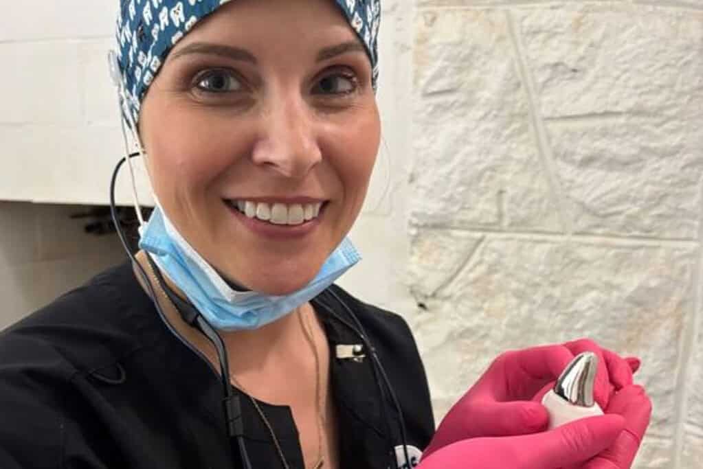Dental professional wearing protective gear, smiling, holding dental tools in a clinical setting.