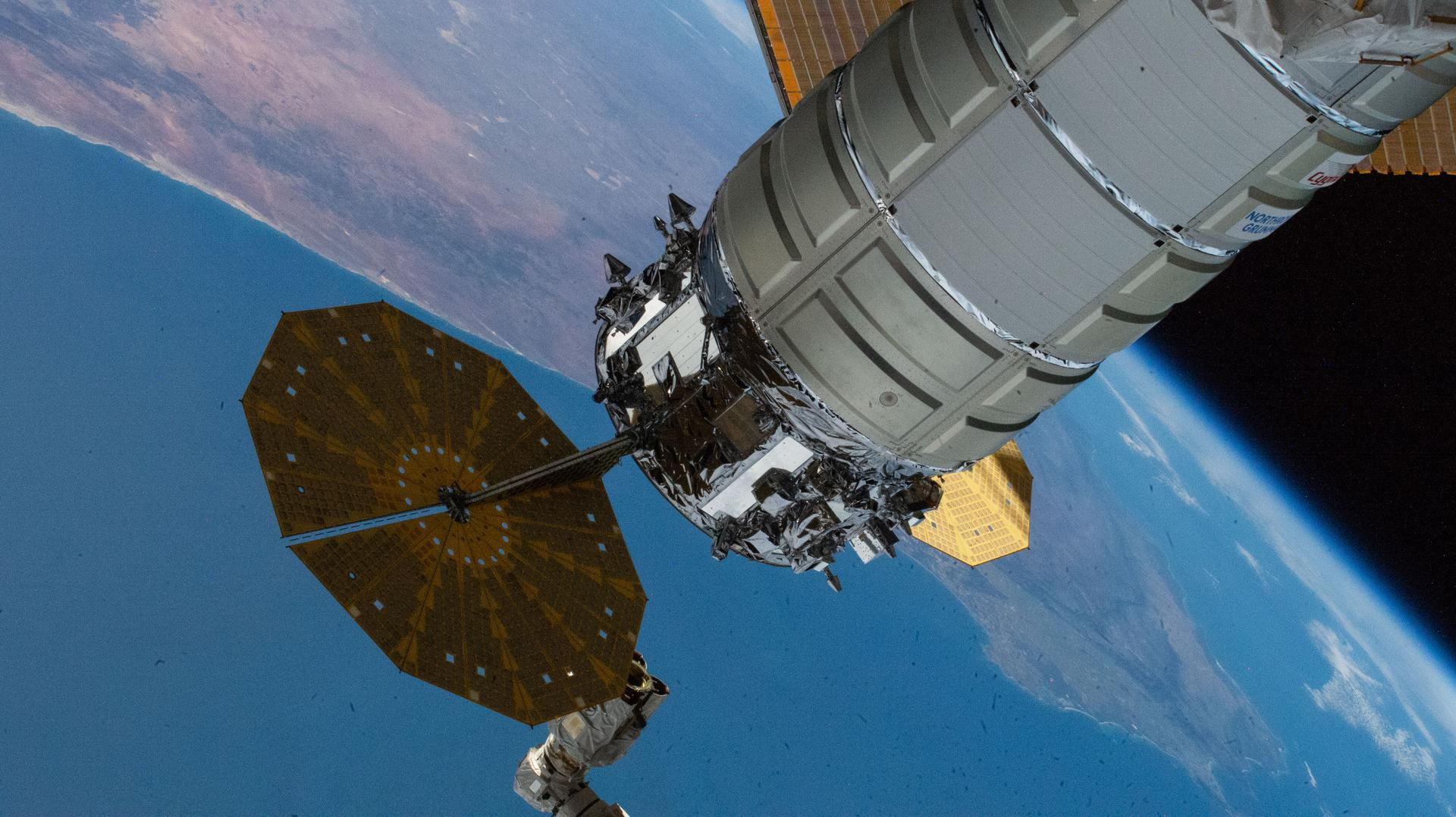 a cylindrical cargo spacecraft is seen attached to the international space station, with earth in the background