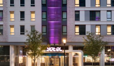 Modern hotel entrance with revolving glass doors, illuminated Yotel sign, streetlamp, trees, and light trails from passing traffic at night.