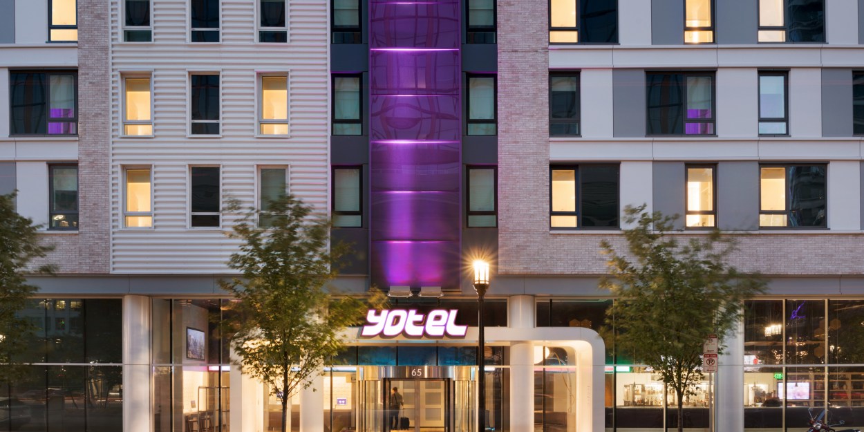 Modern hotel entrance with revolving glass doors, illuminated Yotel sign, streetlamp, trees, and light trails from passing traffic at night.