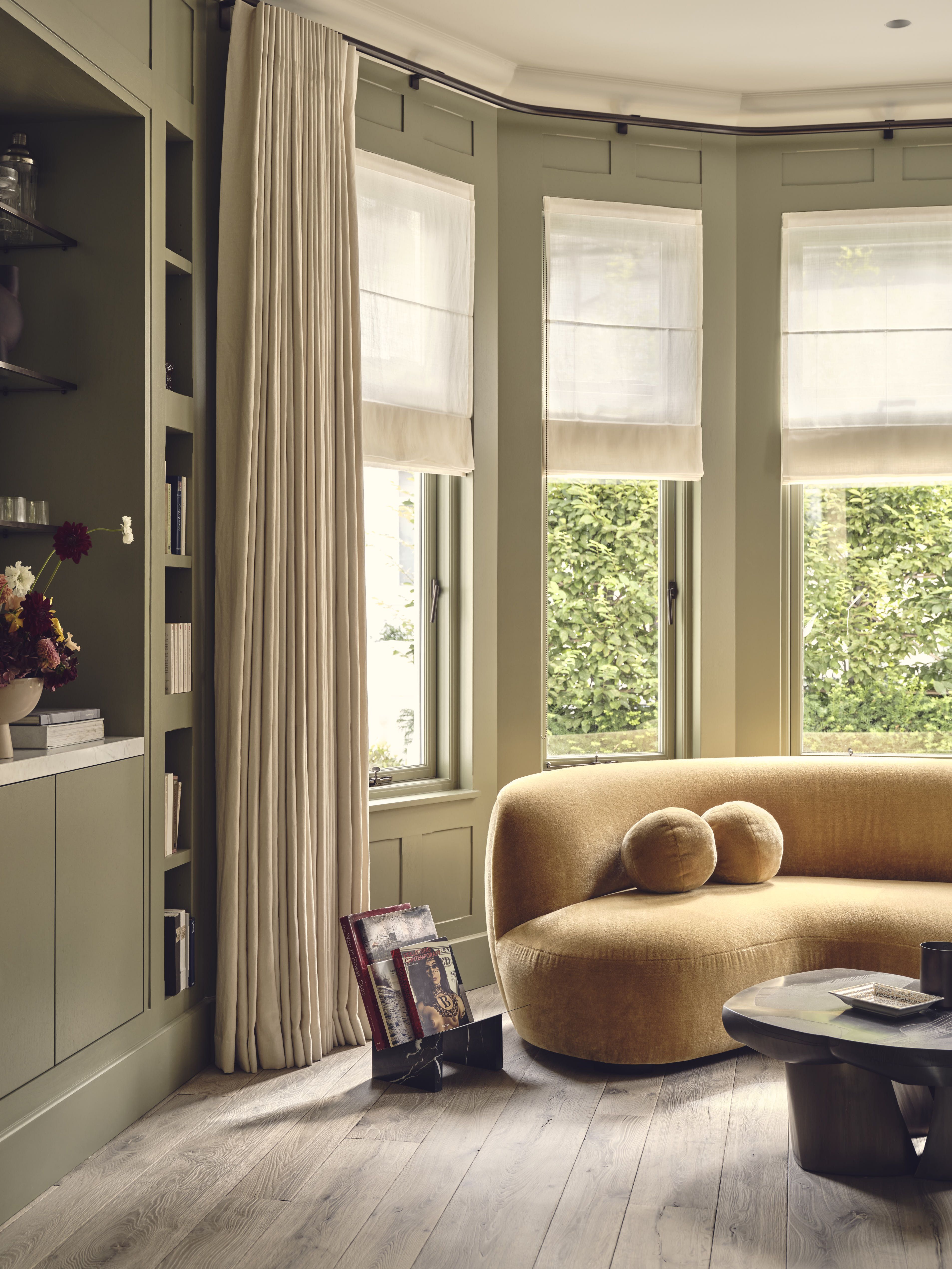 modern living room with sheer roman blinds layered with curtains, green joinery and walls, curved yellow velvet sofa with magazine rack and timber coffee table