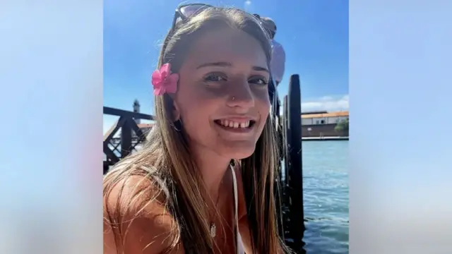 Family photo of 18-year-old Juliette Kenny. Shes sitting on a wooden dock on the canal in Venice. She's wearing a white spaghetti strap top and a pink flower over her right ear