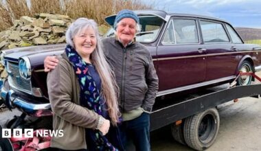 A woman with long grey hair stands next to a man with a blue woollen hat. They are standing in front of an old brown car, which is on the back of a lorry.