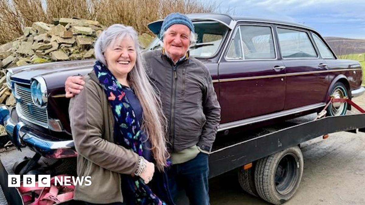A woman with long grey hair stands next to a man with a blue woollen hat. They are standing in front of an old brown car, which is on the back of a lorry.
