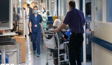 A man leans over an elderly patient lying on a trolley in a hospital corridor, with other patients and equipment visible in the background.