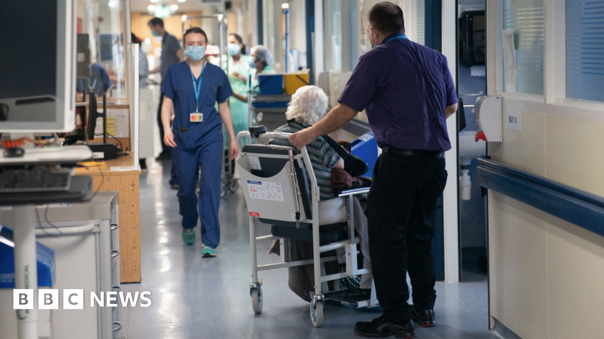 A man leans over an elderly patient lying on a trolley in a hospital corridor, with other patients and equipment visible in the background.