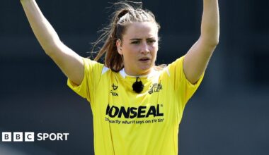 Tara Jones, wearing a blue patterned blazer and pink headpiece, holds up her MBE medal with Windsor Castle in the background