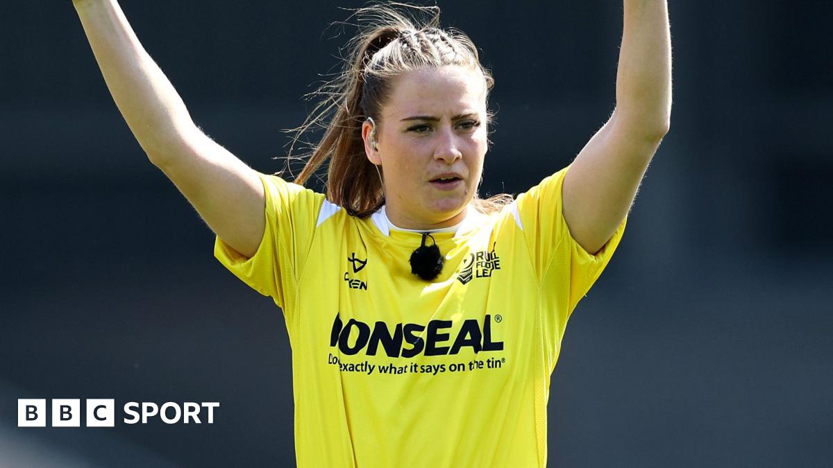 Tara Jones, wearing a blue patterned blazer and pink headpiece, holds up her MBE medal with Windsor Castle in the background