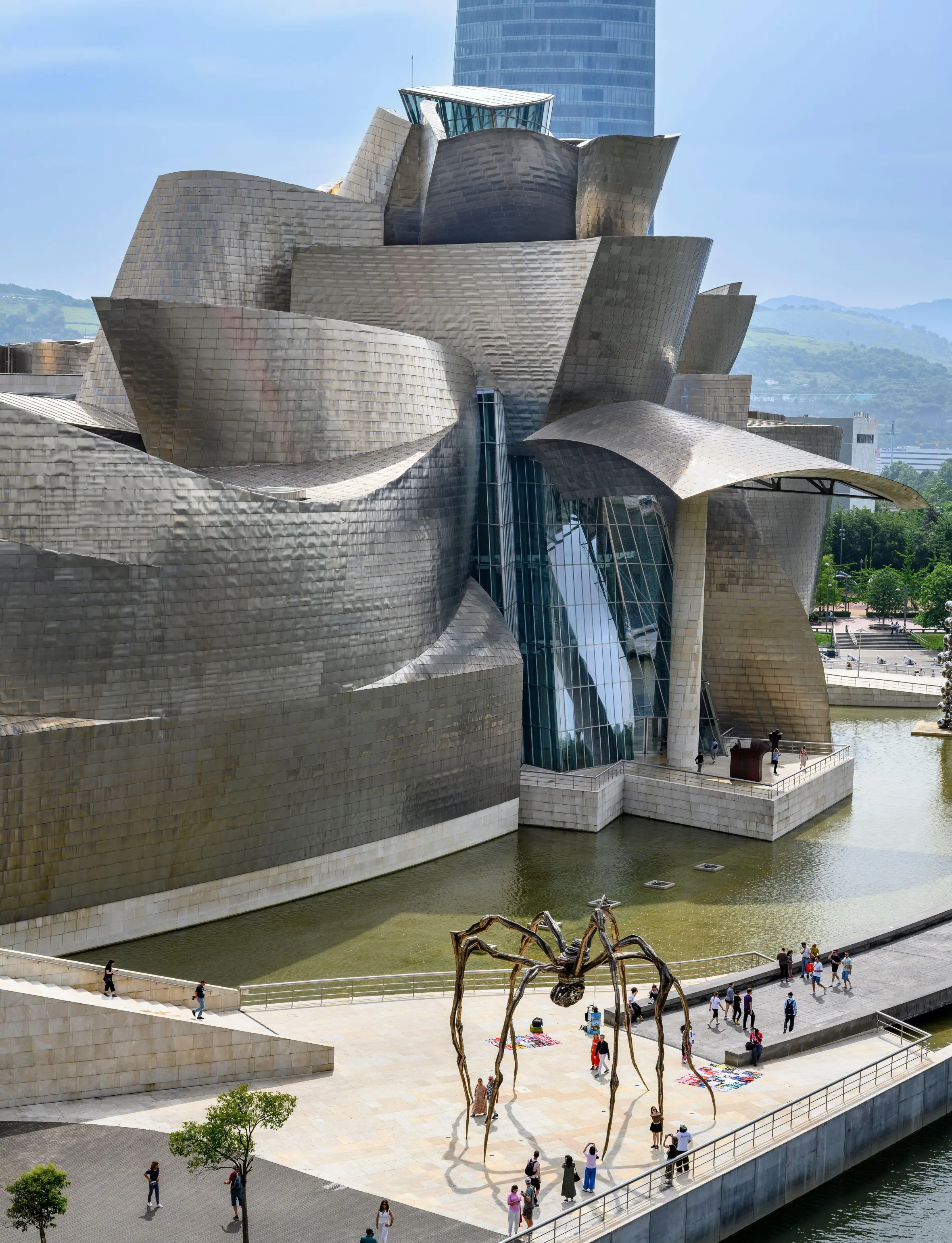 Exterior view of the Guggenheim Museum, Bilbao, featuring its metallic, undulating architecture alongside the Nervion River and a large spider sculpture.
