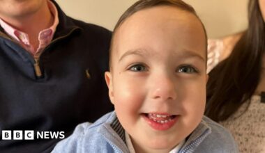 A bright-eyed little boy smiles at the camera. he wears a pale blue jumper. Behind him are sat his parents.