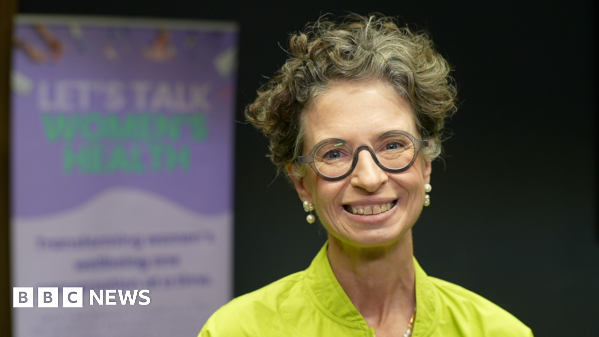 Dr Liz O'Riordan wearing a lime green outfit. She is looking directly at the camera and smiling. Behind her is a purple information board with lettering that says, LET'S TALK WOMEN'S HEALTH.
