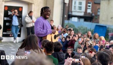 Myles Smith wearing a purple jumper and singing while playing guitar to a gathered crowd on a Luton street.
