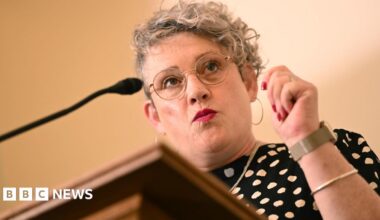 Ashley Dalton has curly blonde hair and wears a black dress with white polka dots. She is standing in front of a microphone at a lectern.