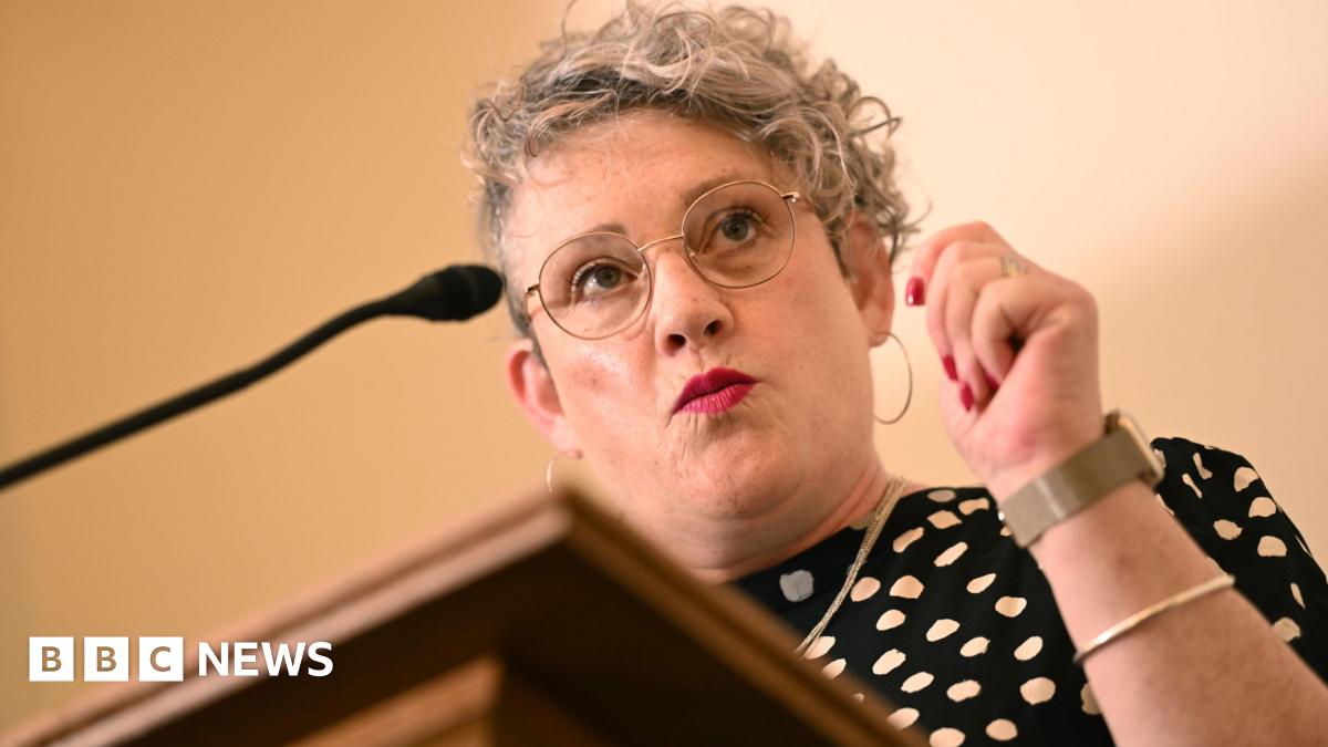 Ashley Dalton has curly blonde hair and wears a black dress with white polka dots. She is standing in front of a microphone at a lectern.