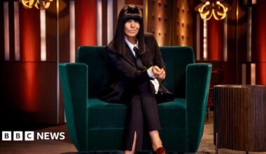 Claudia Winkleman sits on a green velvet sofa wearing a burgundy blazer and trousers