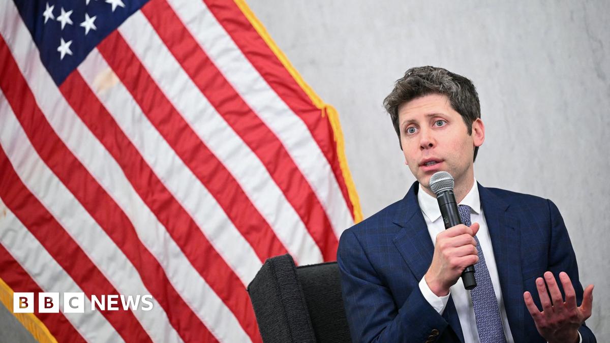 The OpenAI CEO Sam Altman is wearing a dark blue suit with a white shirt and blue and white tie. He is holding and speaking into a microphone while sitting in front of the American flag
