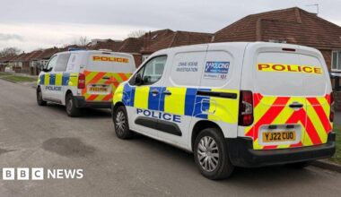 The image shows a residential street with low, single‑storey houses on either side. Two marked police vans are parked along the road.