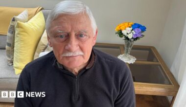 Emlyn Williams in his living room looking at the camera - he's in his 70s and has white hair and a white moustache, and is wearing a black fleece top.  A sofe and a coffee table with a vase of roses are in the background
