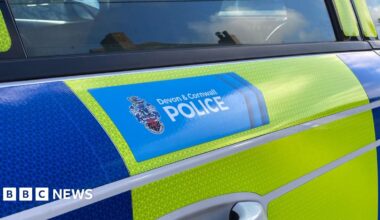 The side of a yellow and blue police vehicle. There is a blue section with the words 'Devon and Cornwall Police' inside of it. The door handle is below and a window above the section.